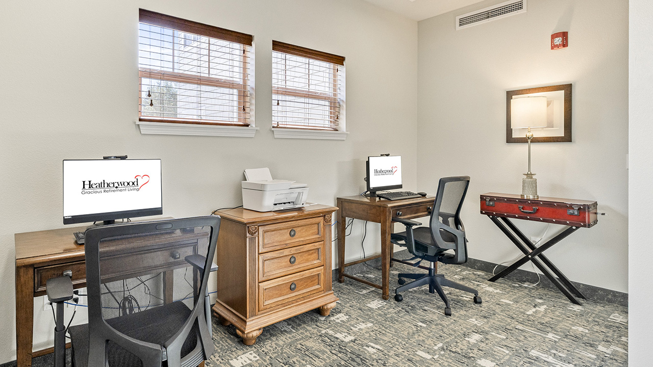 A small office space with two desks, each with a computer and office chair. A printer sits on a wooden cabinet between the desks. Two windows and a lamp on a side table light the room.