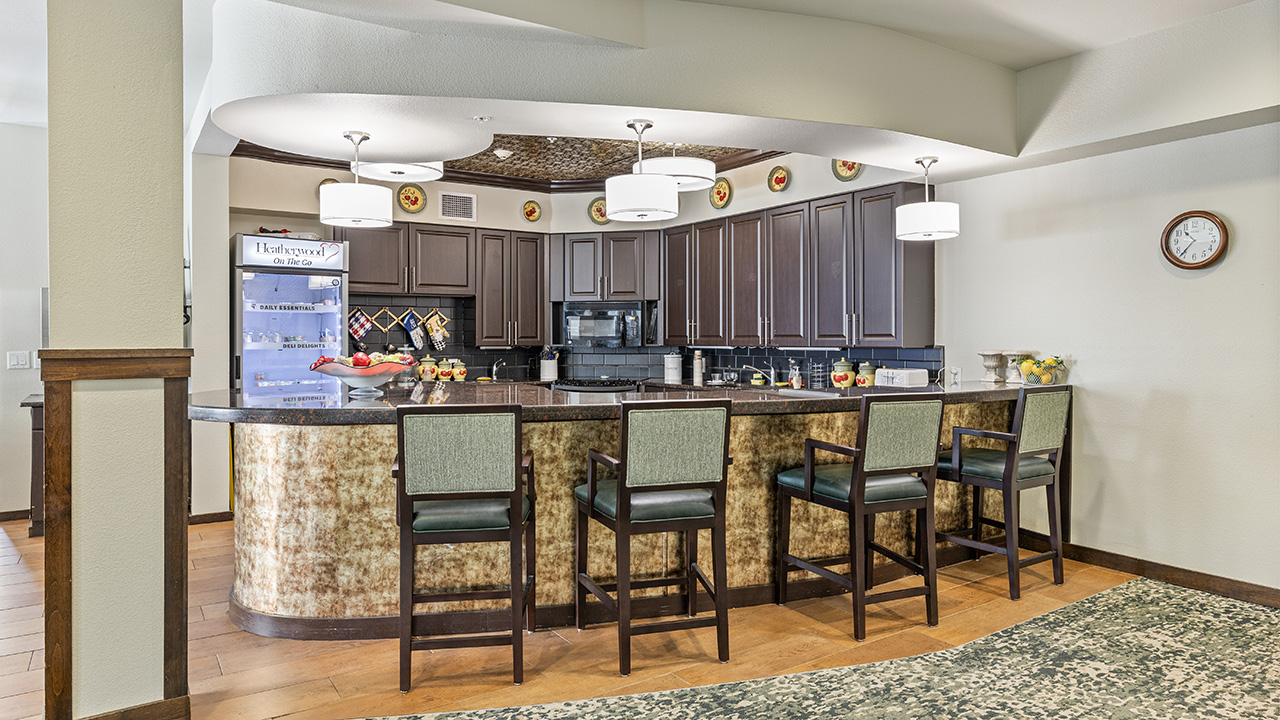A modern kitchen with dark wood cabinets, a long curved countertop, four green-cushioned bar stools, pendant lights, and decorative plates above the cabinets. A clock hangs on the wall to the right.