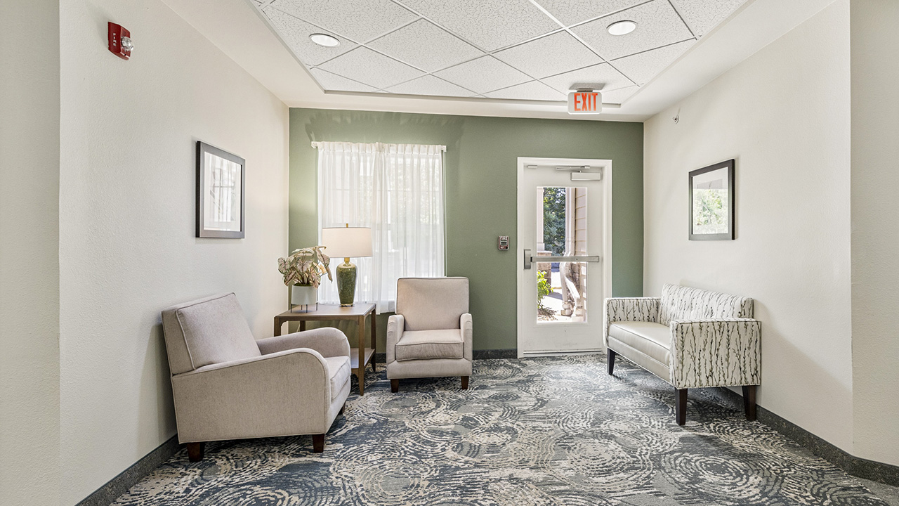 A small, bright waiting area with two beige chairs, a patterned sofa, a lamp on a side table, framed art on cream walls, and a door with a window letting in natural light.