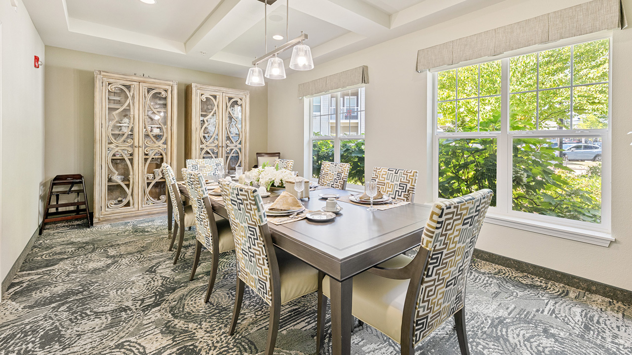A bright dining room with a rectangular table set for eight, patterned chairs, decorative cabinets, large windows letting in natural light, and a modern chandelier overhead.