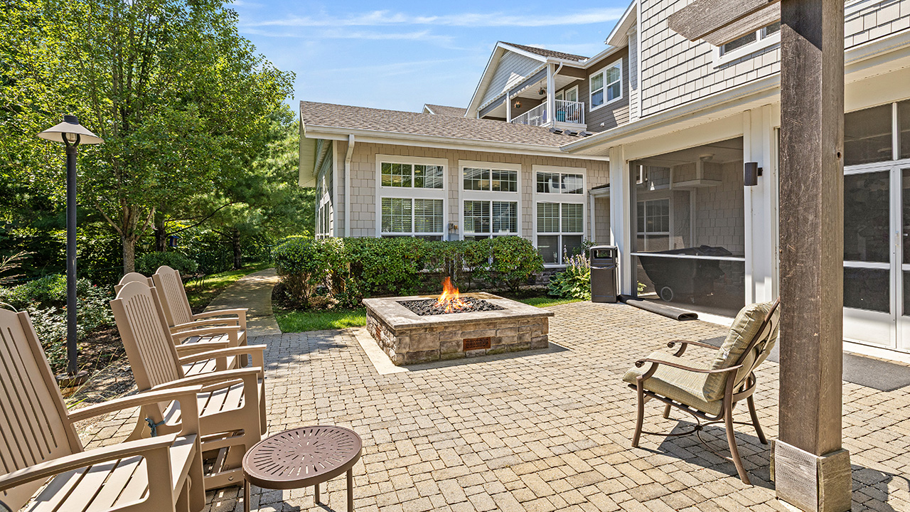 Outdoor patio area with beige chairs arranged around a square stone fire pit, surrounded by greenery and trees, next to a light gray house with large windows under a blue sky.