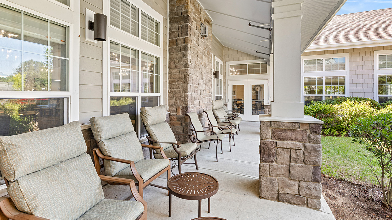 Covered outdoor patio with cushioned chairs and small tables, stone pillars, and large windows. The area overlooks a green lawn with shrubs and is attached to a building with beige siding and stone accents.