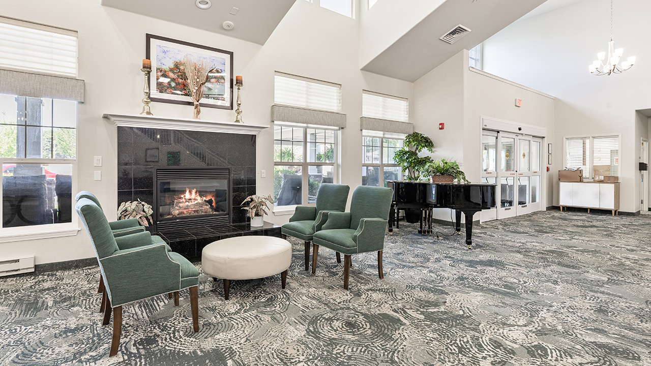 A spacious, well-lit lounge with green armchairs around a white ottoman, a lit fireplace, a grand piano, and large windows letting in natural light. The decor is modern with patterned carpets and plants.