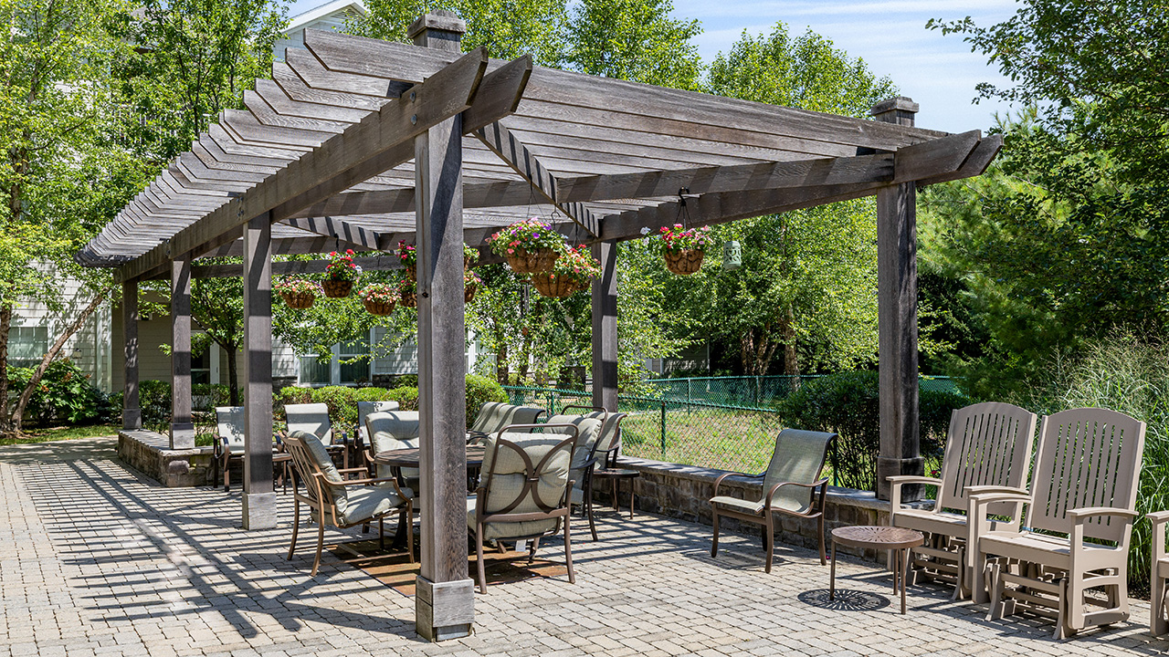 A wooden pergola shades a patio with a dining table and cushioned chairs, surrounded by hanging flower baskets and extra chairs, set in a green, tree-filled outdoor area.