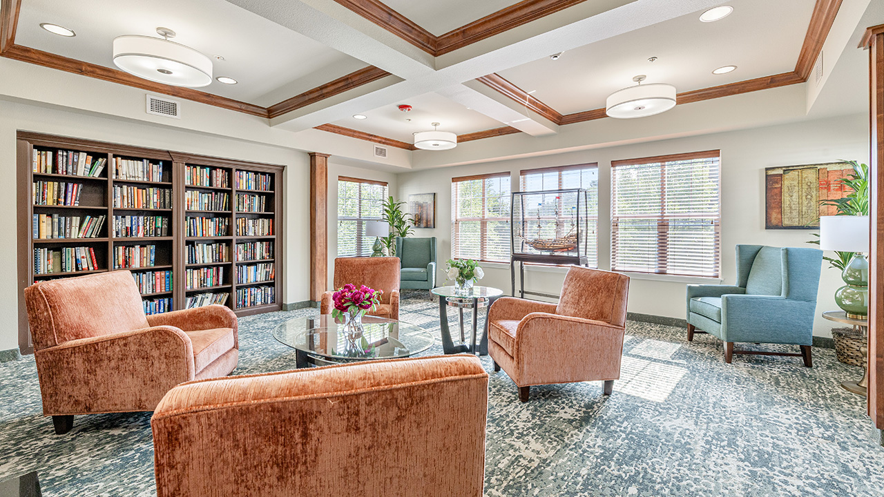 A bright, inviting lounge with four orange armchairs around a glass coffee table, bookshelves along one wall, two blue chairs by large windows, and decorative plants and artwork throughout the room.