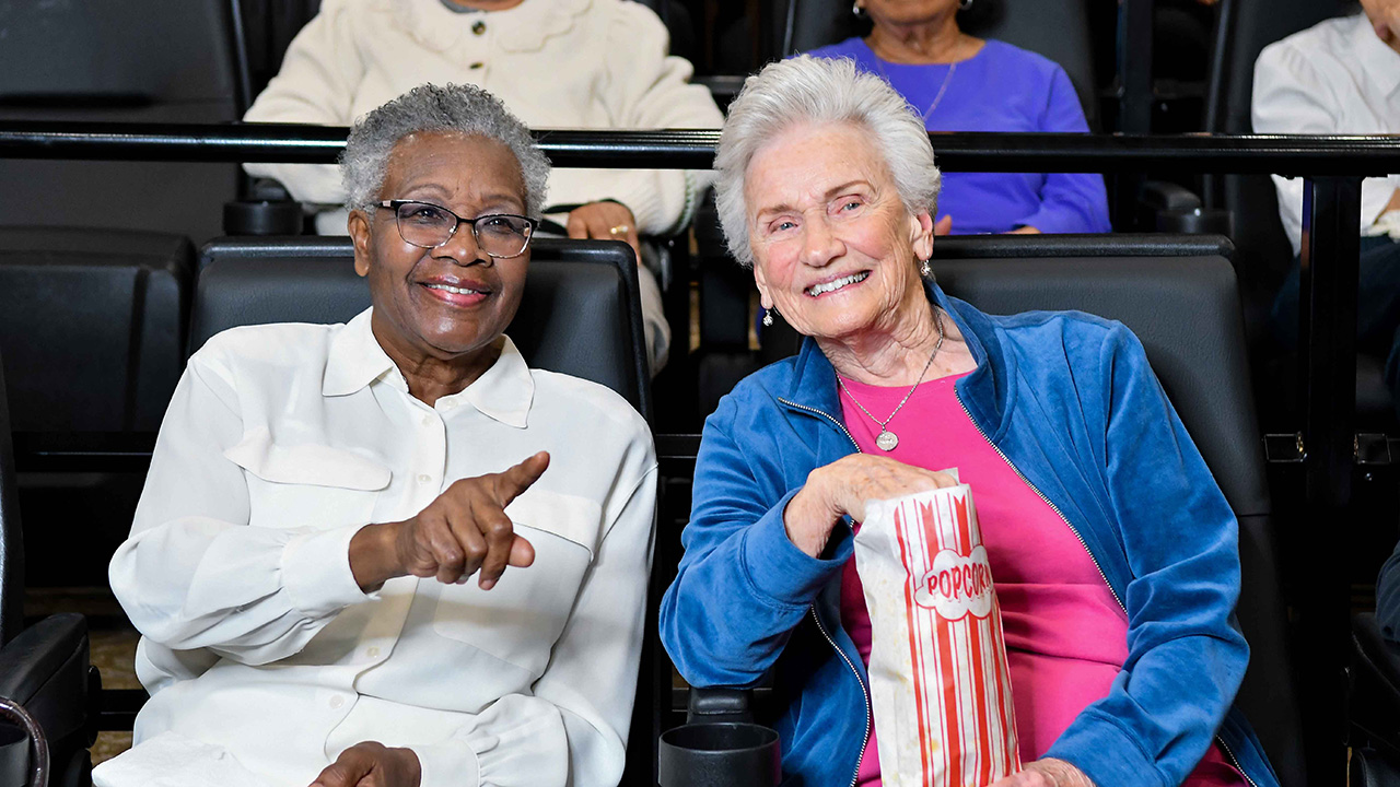 Two smiling older women sit in theater seats; one is pointing ahead and the other holds a striped popcorn bag on her lap. Both appear to be enjoying the movie. Other people are visible sitting behind them.