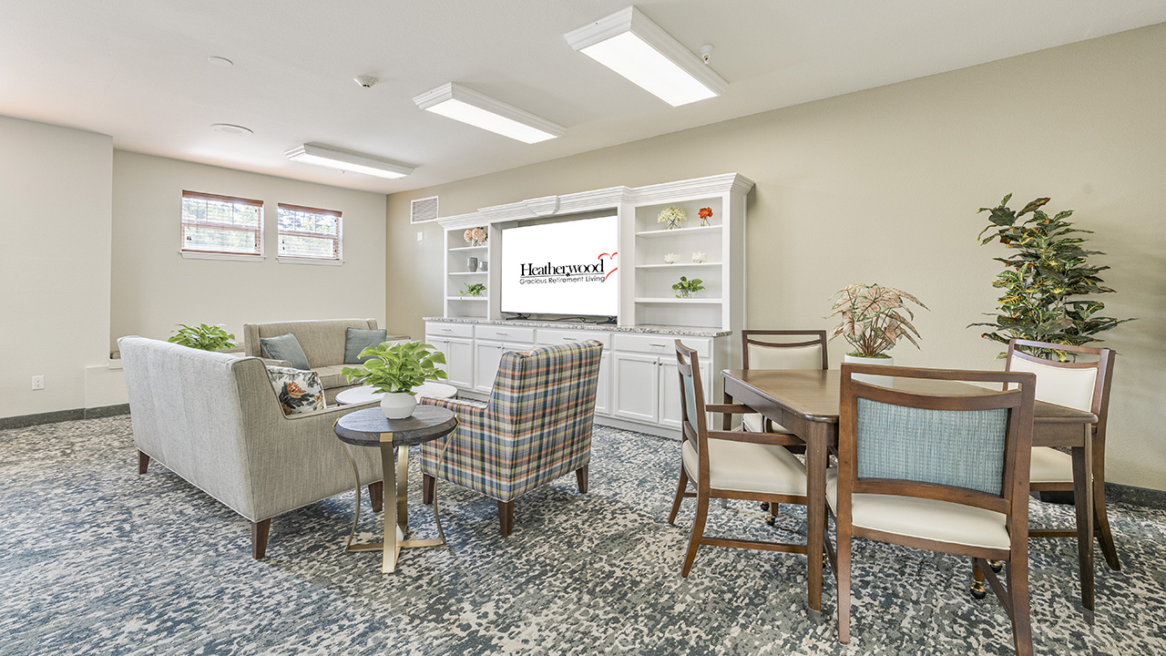 A bright, modern living room with a TV, gray sofa, plaid armchair, wooden dining table with chairs, plants, and white shelves against a beige wall. Natural light streams in from two windows.