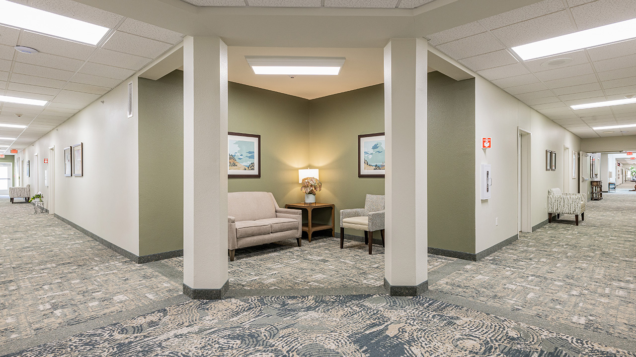 A well-lit hallway in a building features a small seating nook with two beige chairs, a lamp, and framed artwork. The carpet has a patterned design and the walls are painted green and off-white.
