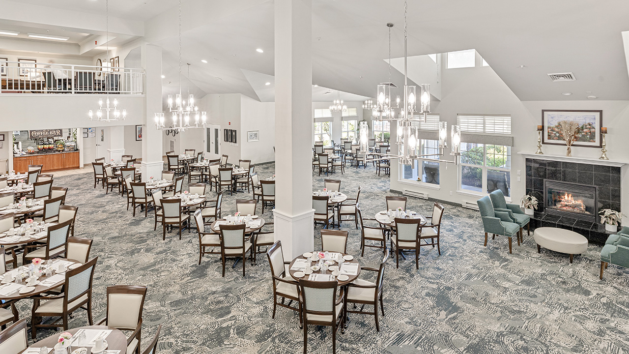 Spacious, elegant dining room with multiple round tables and chairs, set for meals. Large windows, chandeliers, a fireplace with two armchairs, and a balcony area are visible. The atmosphere is bright and inviting.