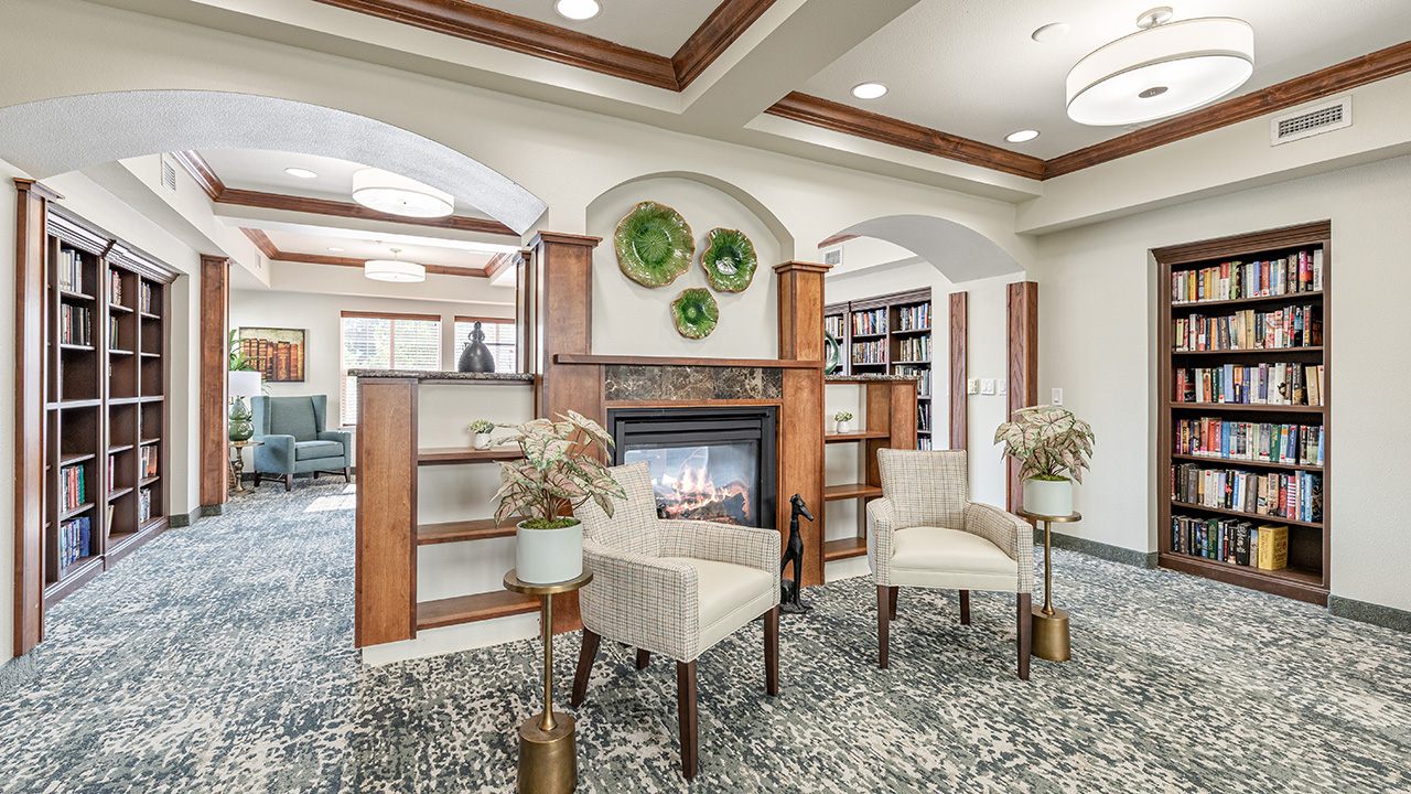 A cozy library with bookshelves lining the walls, a fireplace in the center, two upholstered chairs, decorative plants, and green plates adorning the mantel. Natural light streams in from windows in the background.