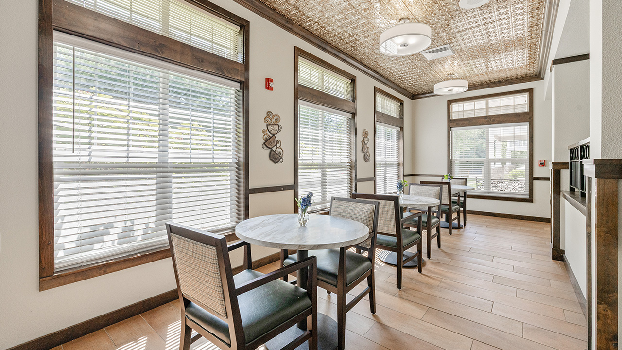 A bright dining area with large windows, round marble-top tables, and wooden chairs. The room has a woven ceiling, tile flooring, and wall decor, creating a cozy and inviting atmosphere.