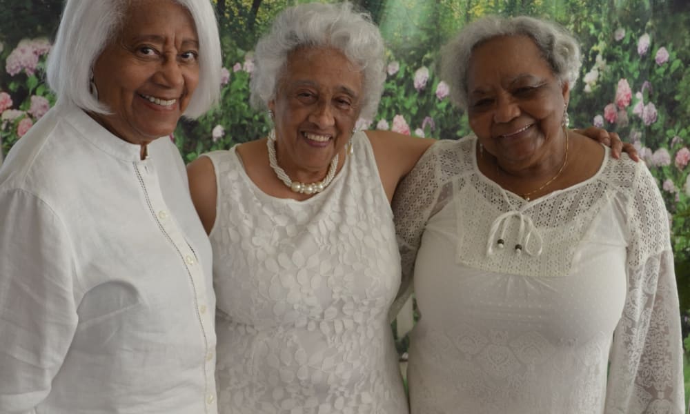 Three elderly women smile warmly, standing close to each other. They are dressed in white clothing, and a floral garden serves as a backdrop. The women appear happy and content, enjoying the moment together.