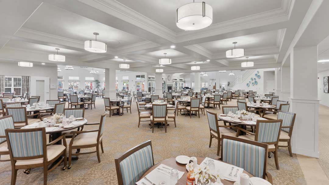 A spacious, well-lit dining area with modern chandeliers, several round tables set for meals, and neatly arranged chairs with light blue cushions. The room features high ceilings and neutral-toned walls, creating an elegant and welcoming atmosphere.