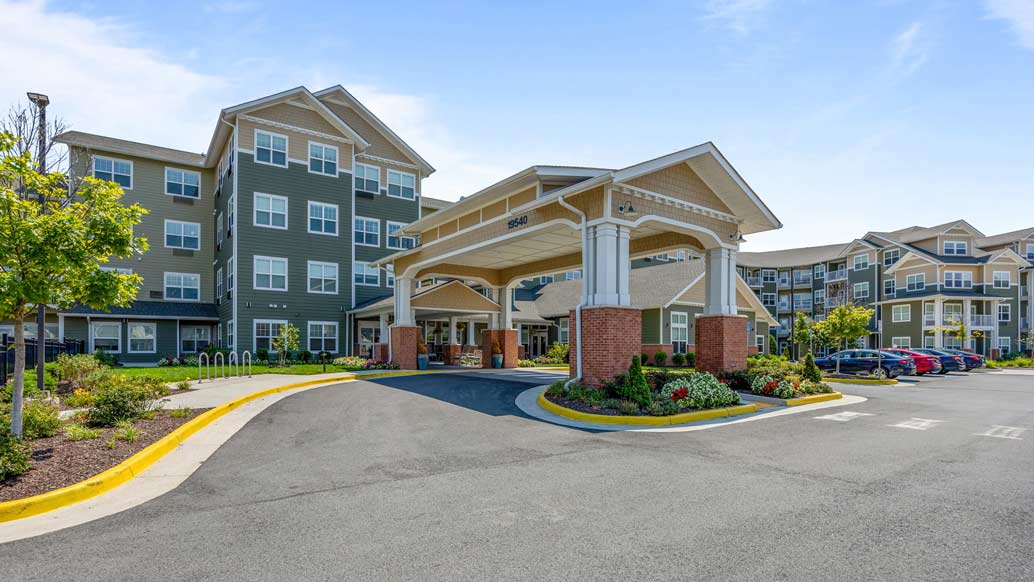 A modern assisted living facility with a large, covered entrance supported by white pillars. The building has multiple stories, green siding, and numerous windows. There are landscaped gardens, a clear blue sky, and parked cars near the entrance.