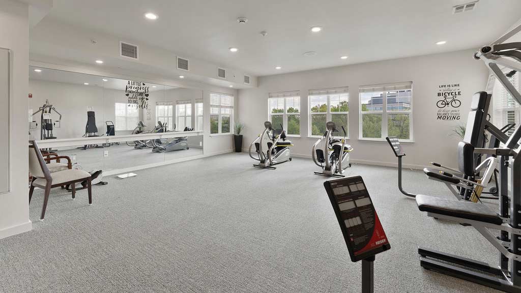 A spacious gym with white walls and carpeted floors featuring various exercise equipment, including two stationary bikes, an elliptical machine, and weight training machines. Large windows allow natural light to brighten the room. Motivational quotes are on the walls.