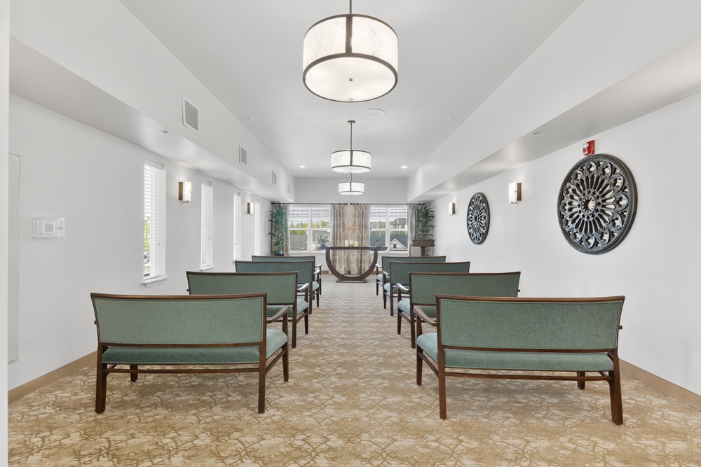 A small, well-lit room with six rows of teal cushioned chairs facing a wooden podium. The walls are white, decorated with two circular art pieces. Large windows at the front let in natural light, and ceiling lights hang in a row.