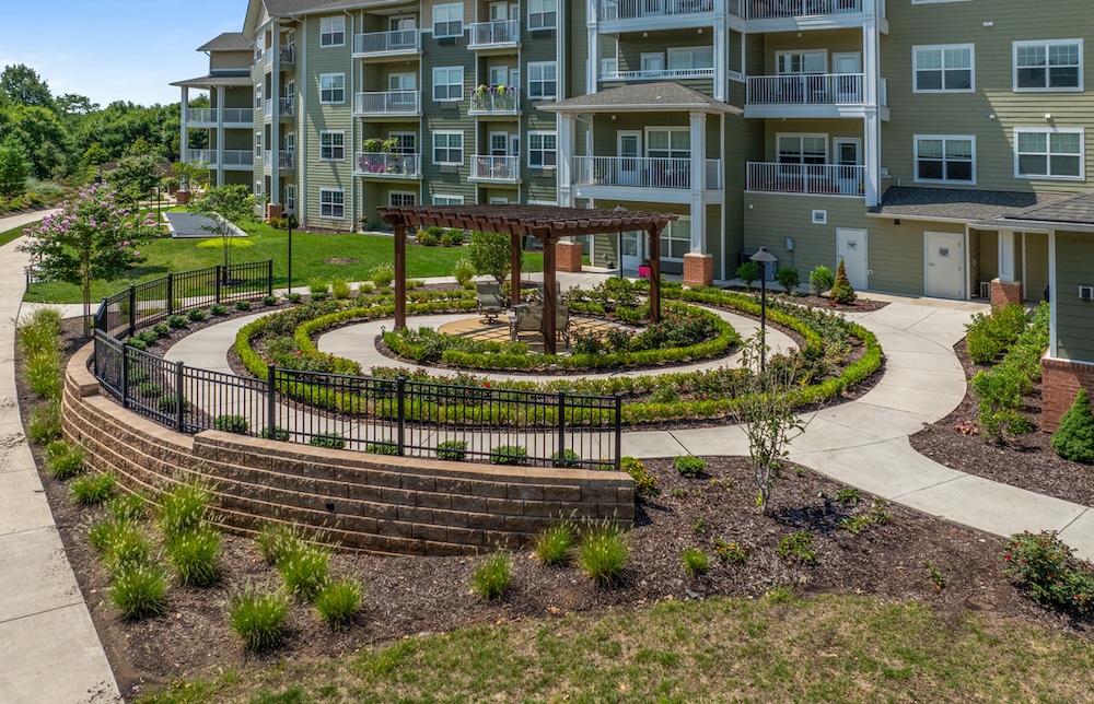 A circular landscaped garden features a central wooden pergola with seating surrounded by pathways and colorful flowers. Behind the garden is a multi-story residential building with balconies.