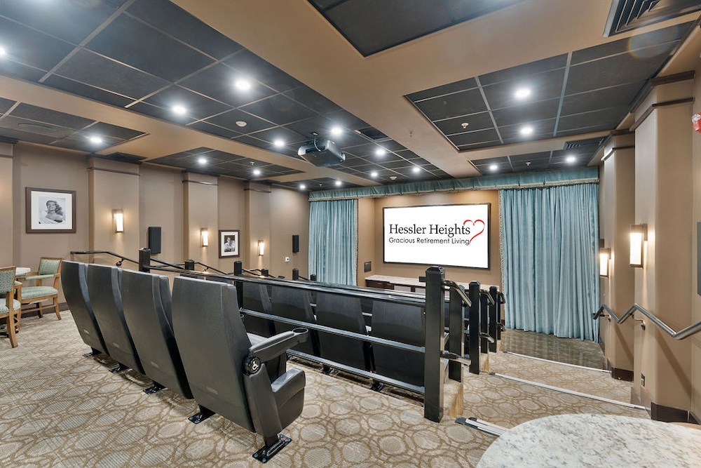 A modern theater room with several rows of black cushioned seats facing a large screen. The screen displays 