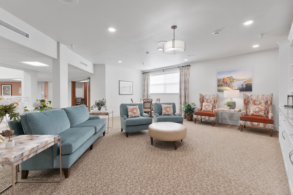 A bright living room with a blue sofa, two matching armchairs, and an ottoman around a patterned rug. Two accent chairs with orange cushions are against the wall, with decorative plants and artwork. Overhead lighting and large windows illuminate the space.