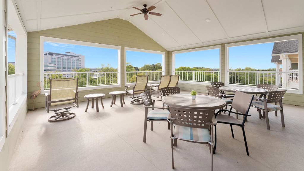 Enclosed patio with large windows offering scenic views. The space features several tables with chairs, comfortable armchairs, and ceiling fans. A mix of beige and brown tones complements the airy, well-lit atmosphere.