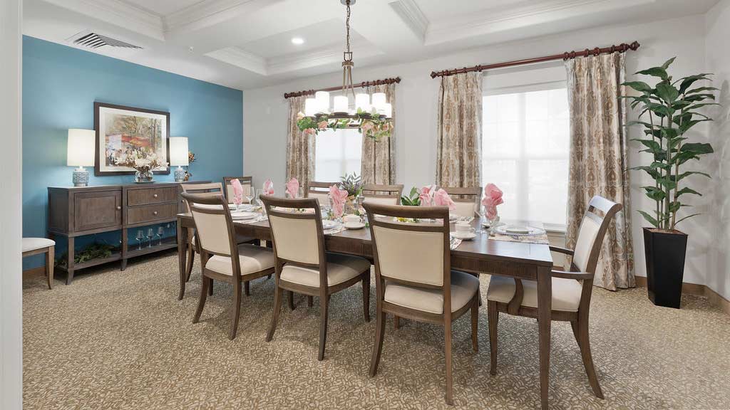 A well-lit dining room with a rectangular wooden table set with pink napkins, plates, and silverware. The room has ten beige cushioned chairs, a blue accent wall with a painting and two lamps on a sideboard, patterned curtains, and a potted plant in the corner.