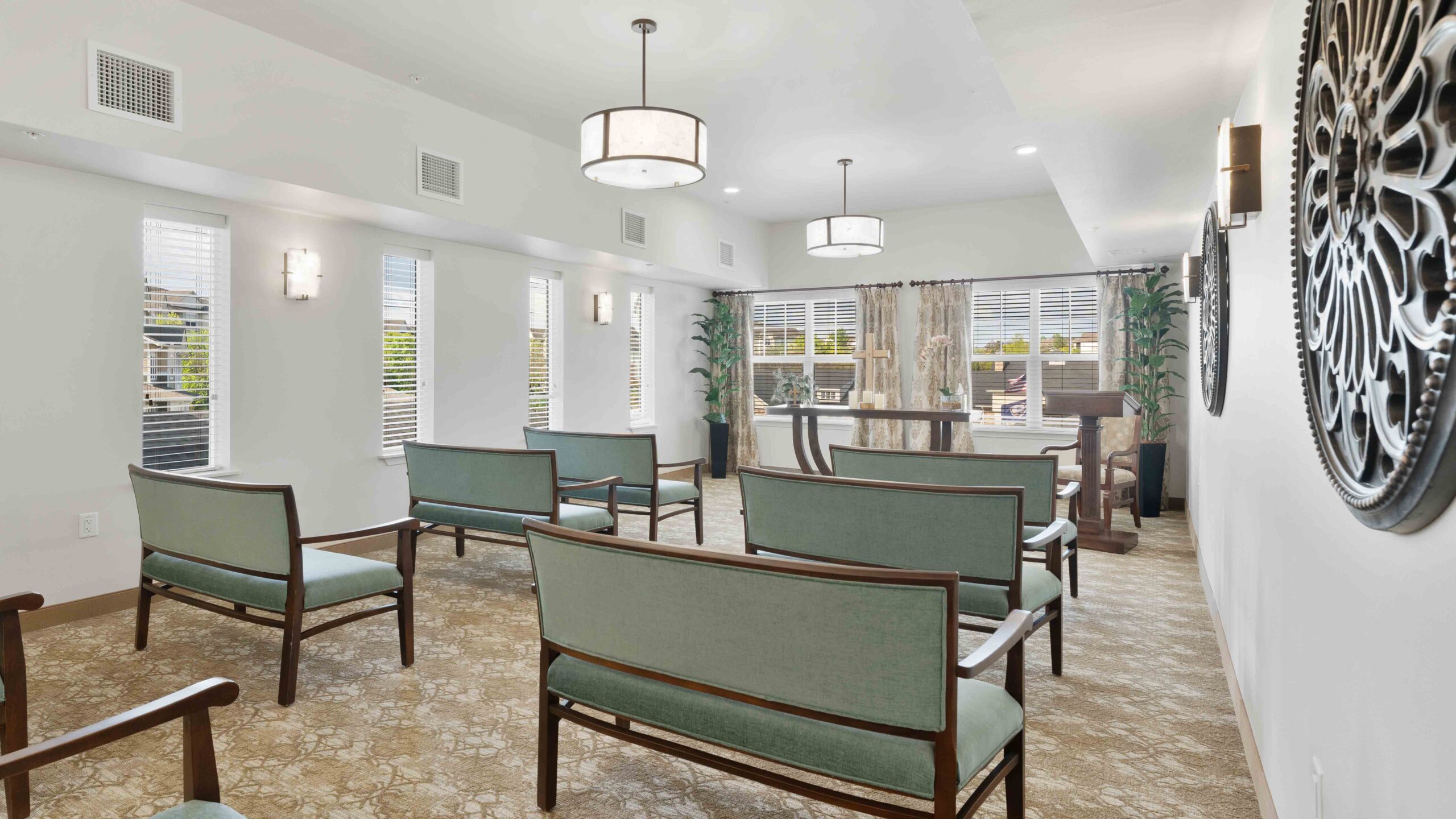 A bright, modern waiting room with green cushioned chairs, large windows with blinds, patterned carpet, a decorative wall piece, plants, and overhead lighting fixtures.