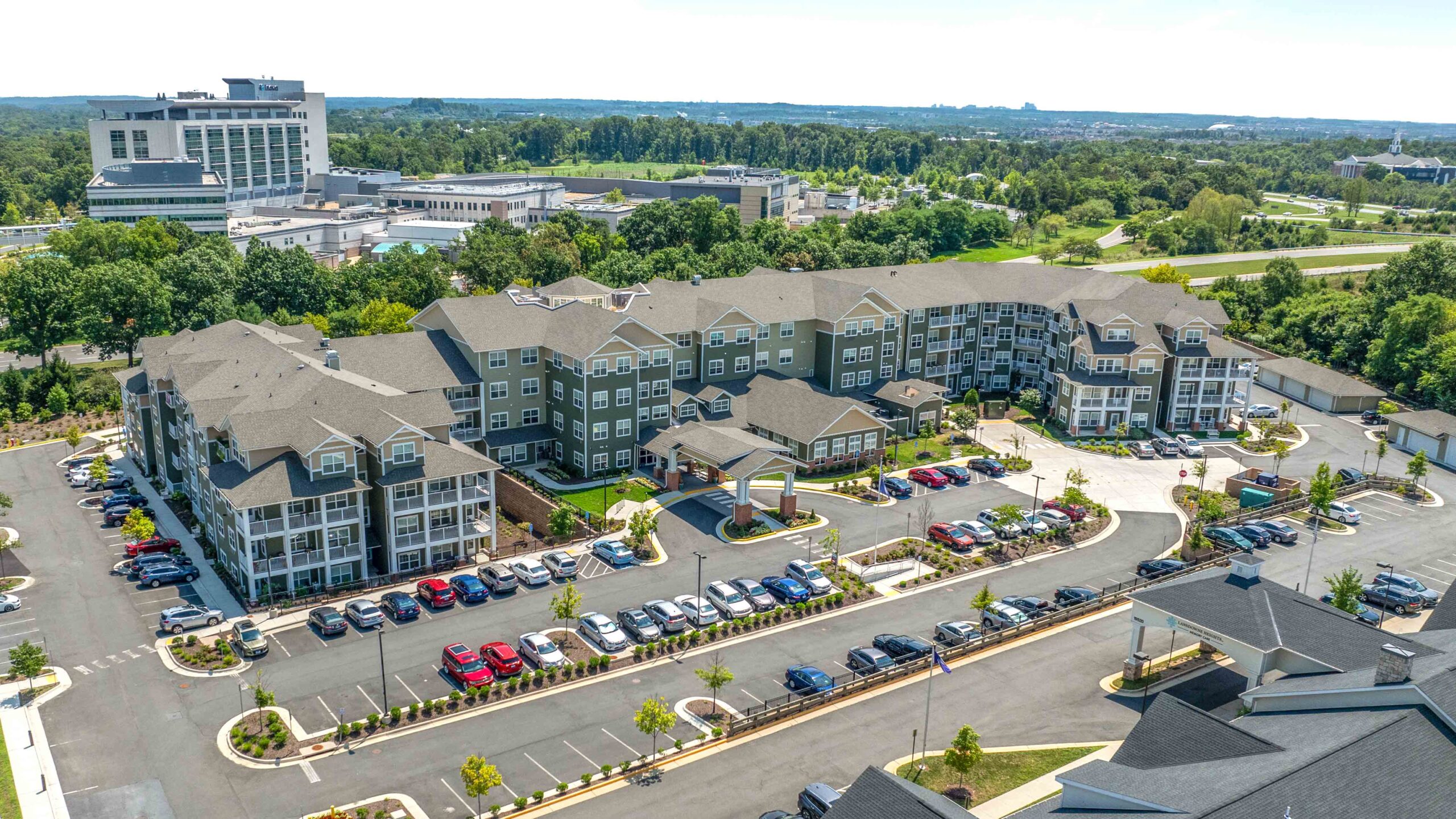Aerial view of a large, modern apartment complex with multiple buildings, landscaped grounds, and parking lots filled with cars. Trees and other buildings are visible in the background.