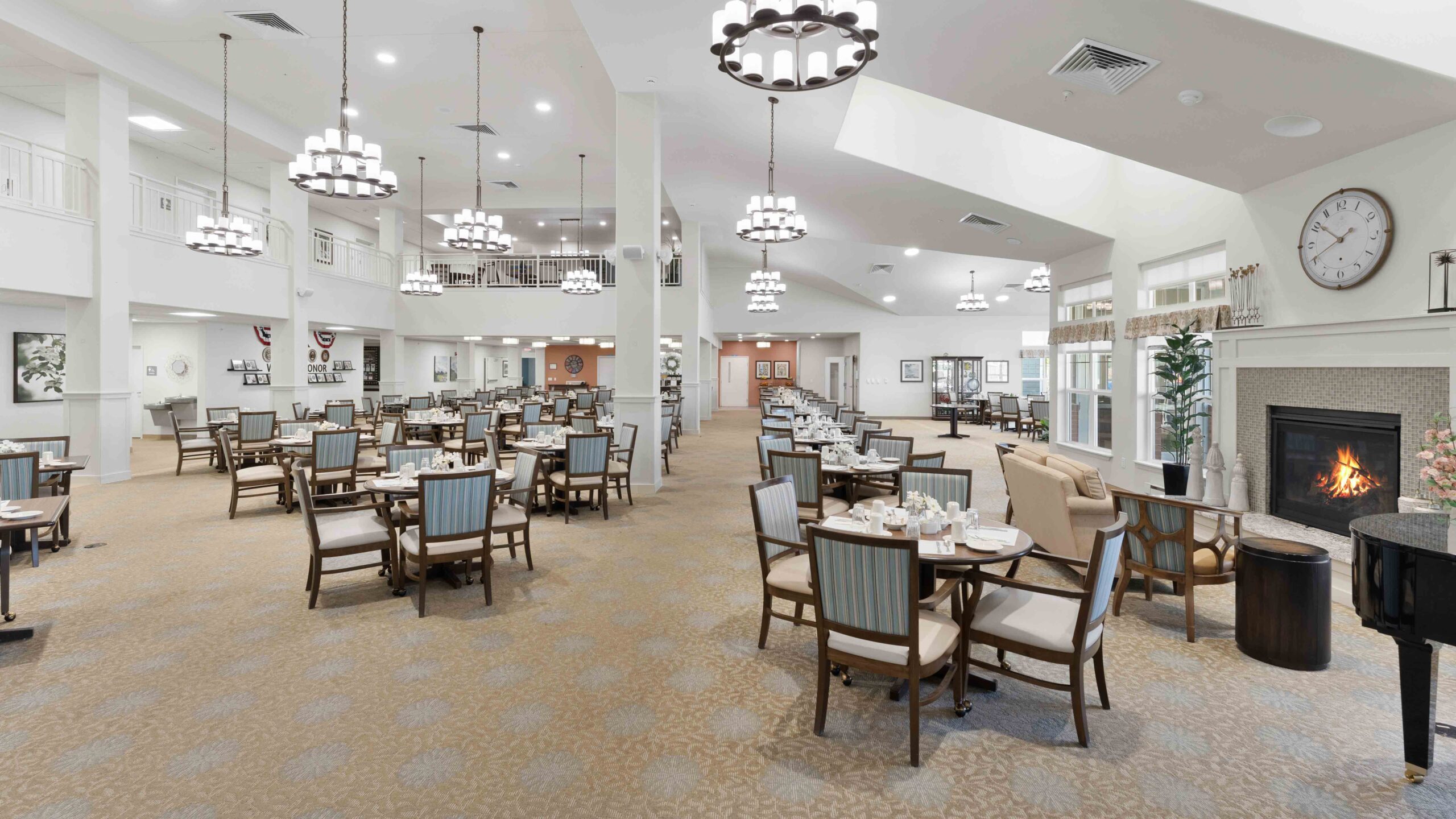 Spacious dining area with neatly arranged tables and chairs, elegant chandeliers, a fireplace, large clock, and bright natural lighting from multiple windows, creating a welcoming and comfortable atmosphere.