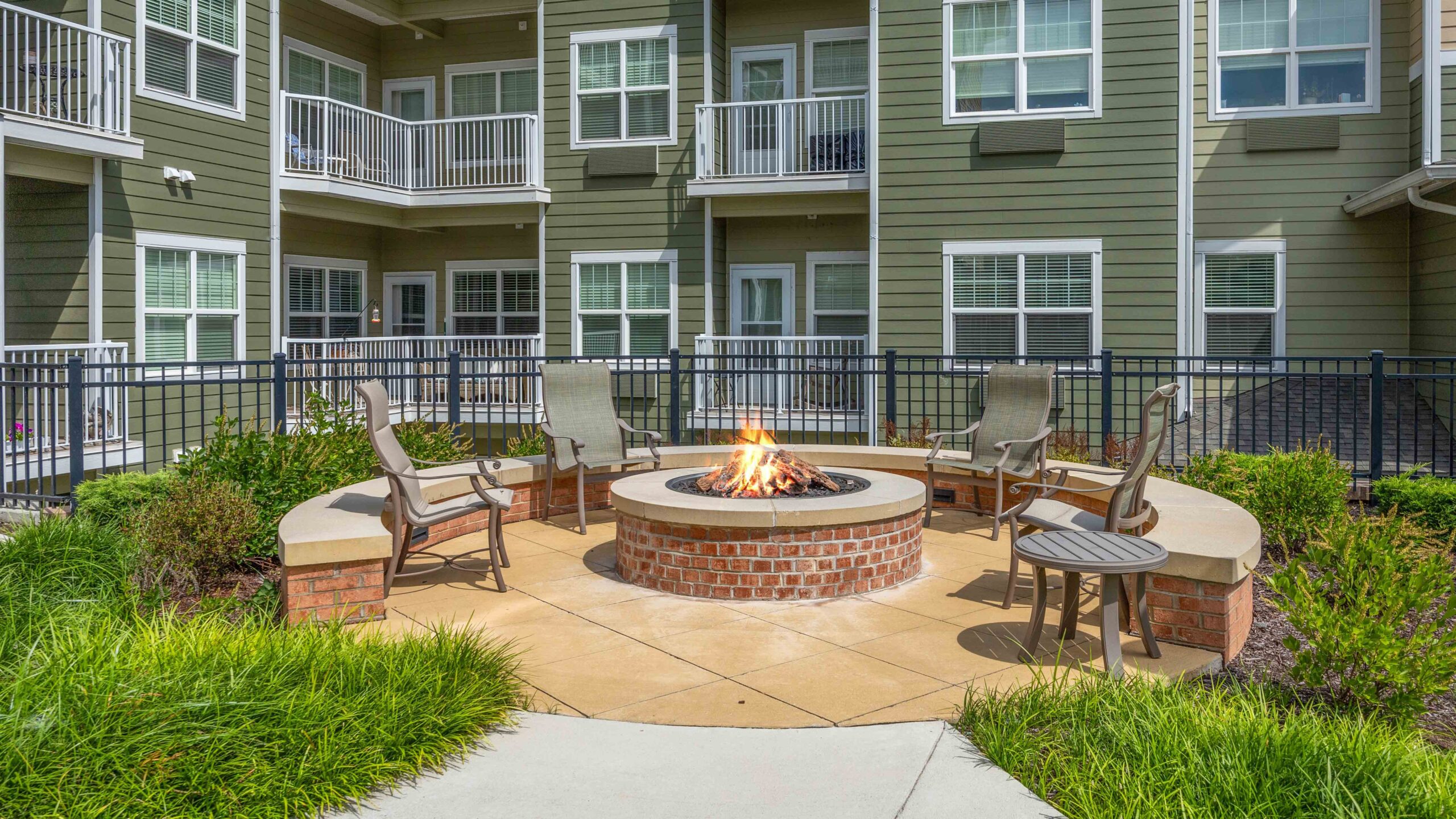Outdoor patio with four chairs around a circular brick fire pit, surrounded by greenery and black metal fencing, in front of a modern apartment building with balconies and large windows.
