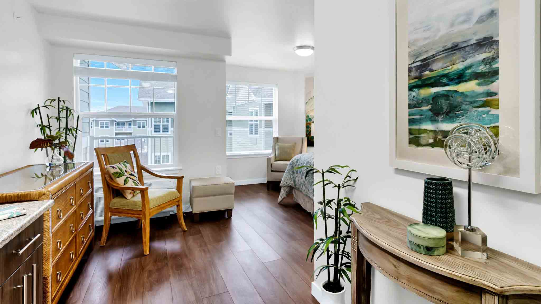 Bright, modern bedroom with wood flooring, large windows, a wooden chair with a cushion, a chest of drawers, green plants, and abstract artwork on white walls. A bed and armchair are visible in the connecting space.