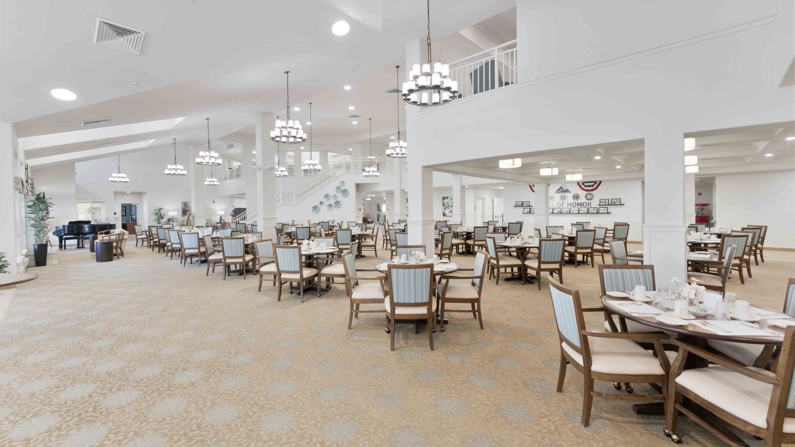 A spacious, well-lit dining room with neatly arranged tables and chairs, light-colored decor, high ceilings, chandeliers, and large windows letting in natural light. The atmosphere is clean and inviting.