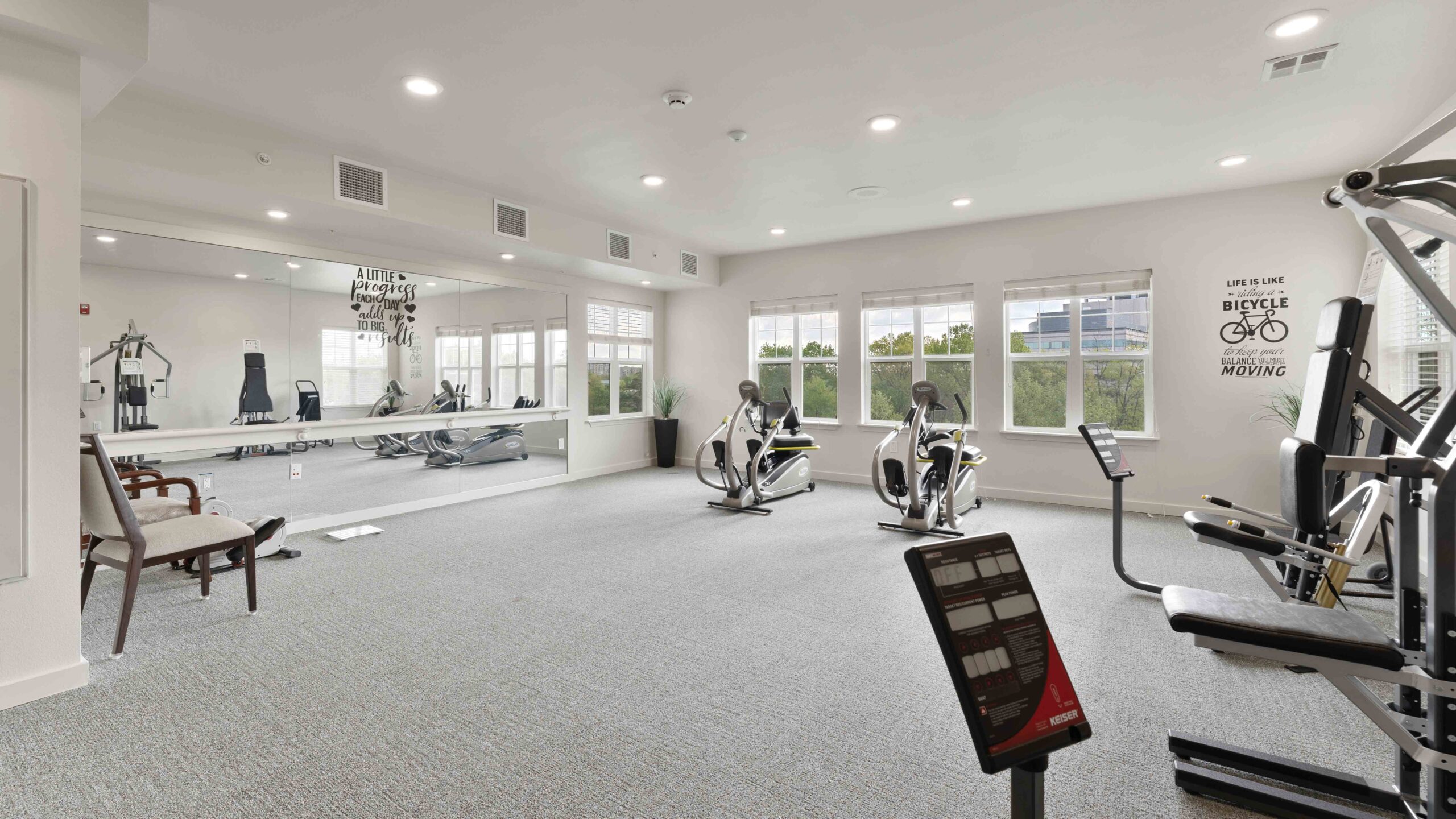 A bright, modern gym with large windows, stationary bikes, strength machines, mirrors, and motivational wall art. The space is clean, spacious, and well-lit with neutral decor and gray carpet.