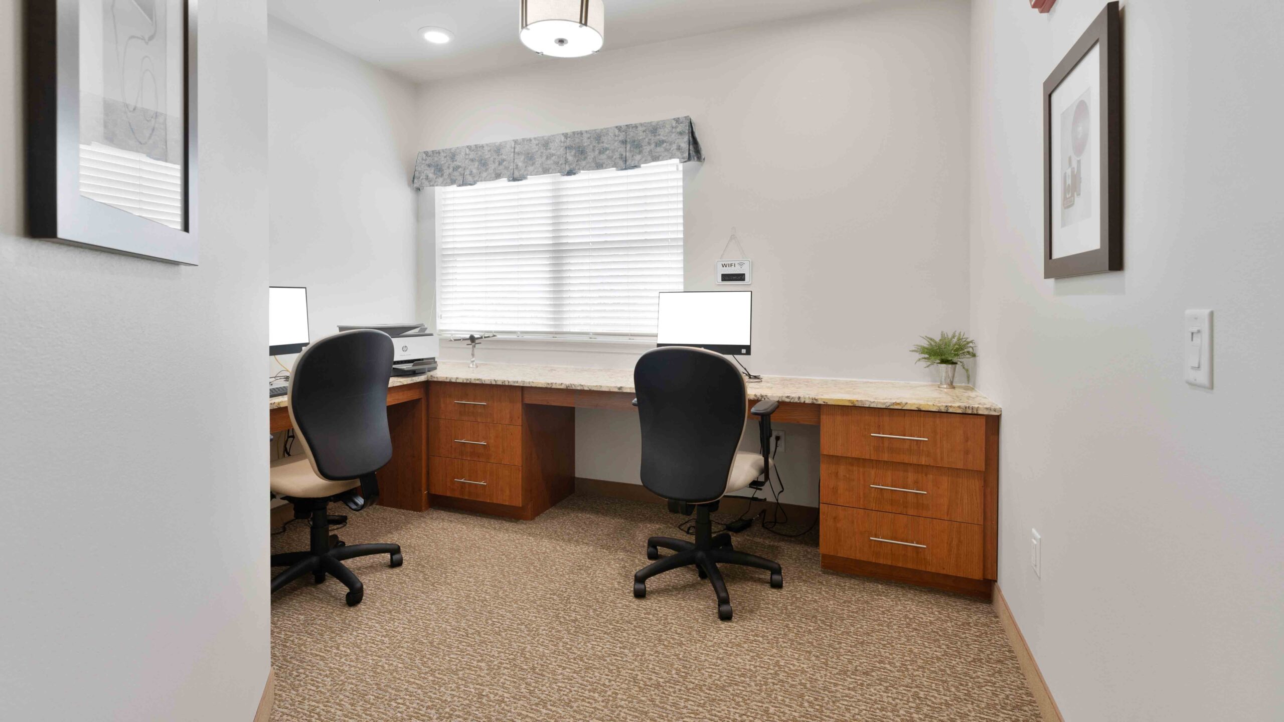 Small office with two desks, each with a computer, chair, and printer under a window with blinds and a valance. The room has beige carpet, artwork on the walls, and a small plant on one desk.