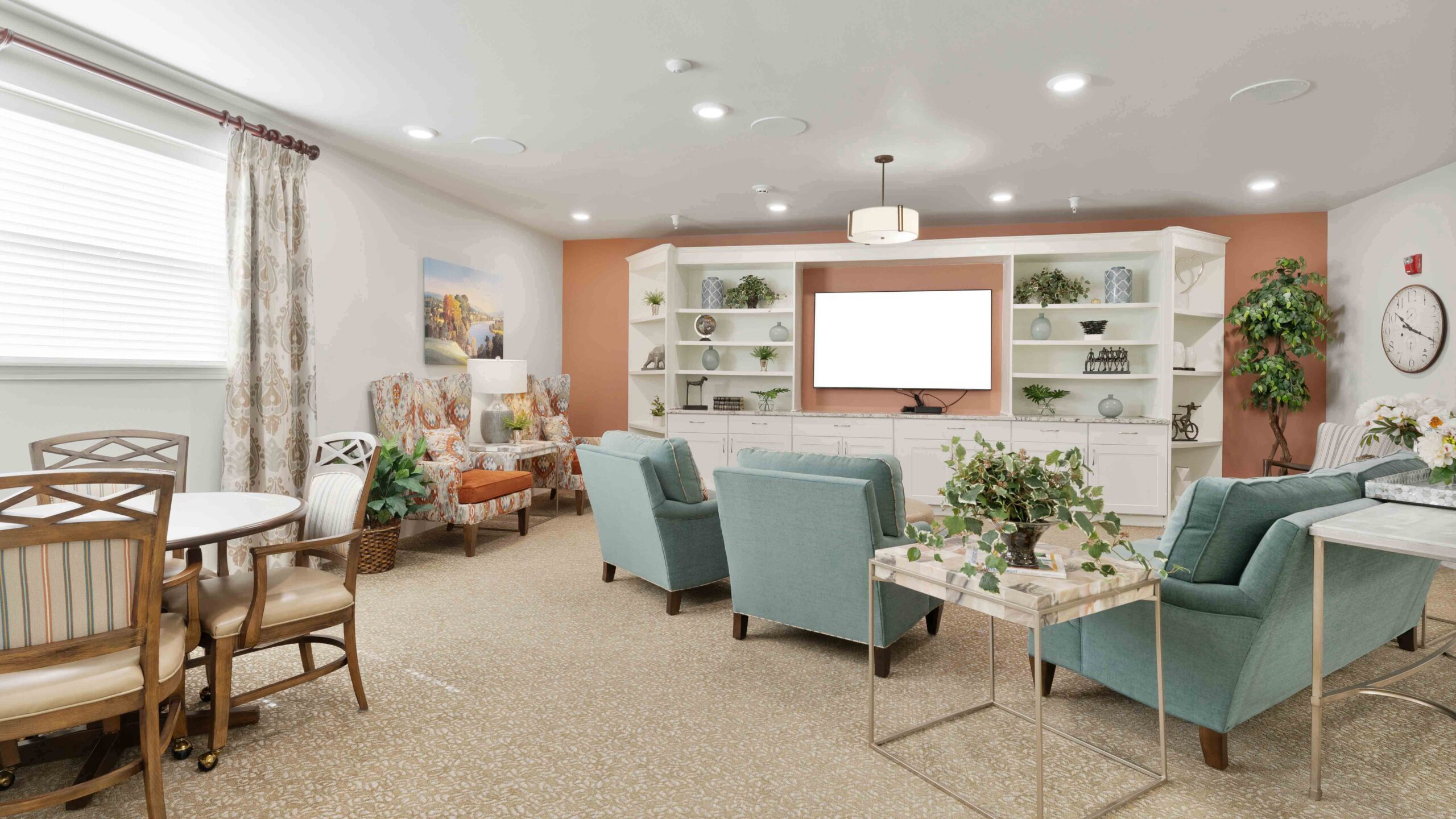 A bright, cozy living room with teal armchairs, a round dining table, built-in white shelves, TV, decorative plants, and large windows with curtains. The space has soft lighting and neutral carpeted flooring.