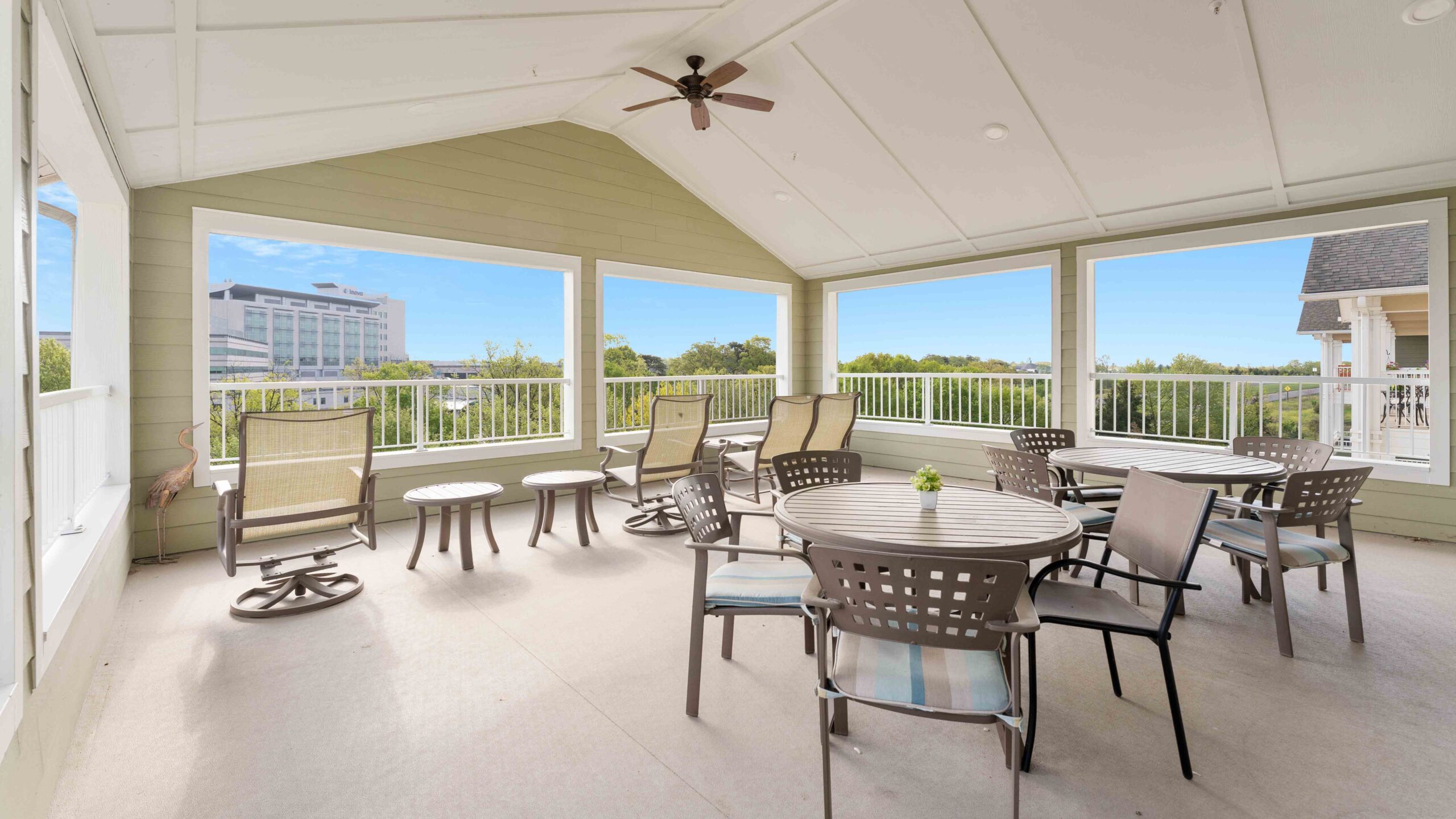 A covered outdoor patio with round tables and chairs, ceiling fan, and large windows providing views of trees and a distant office building under a bright blue sky.