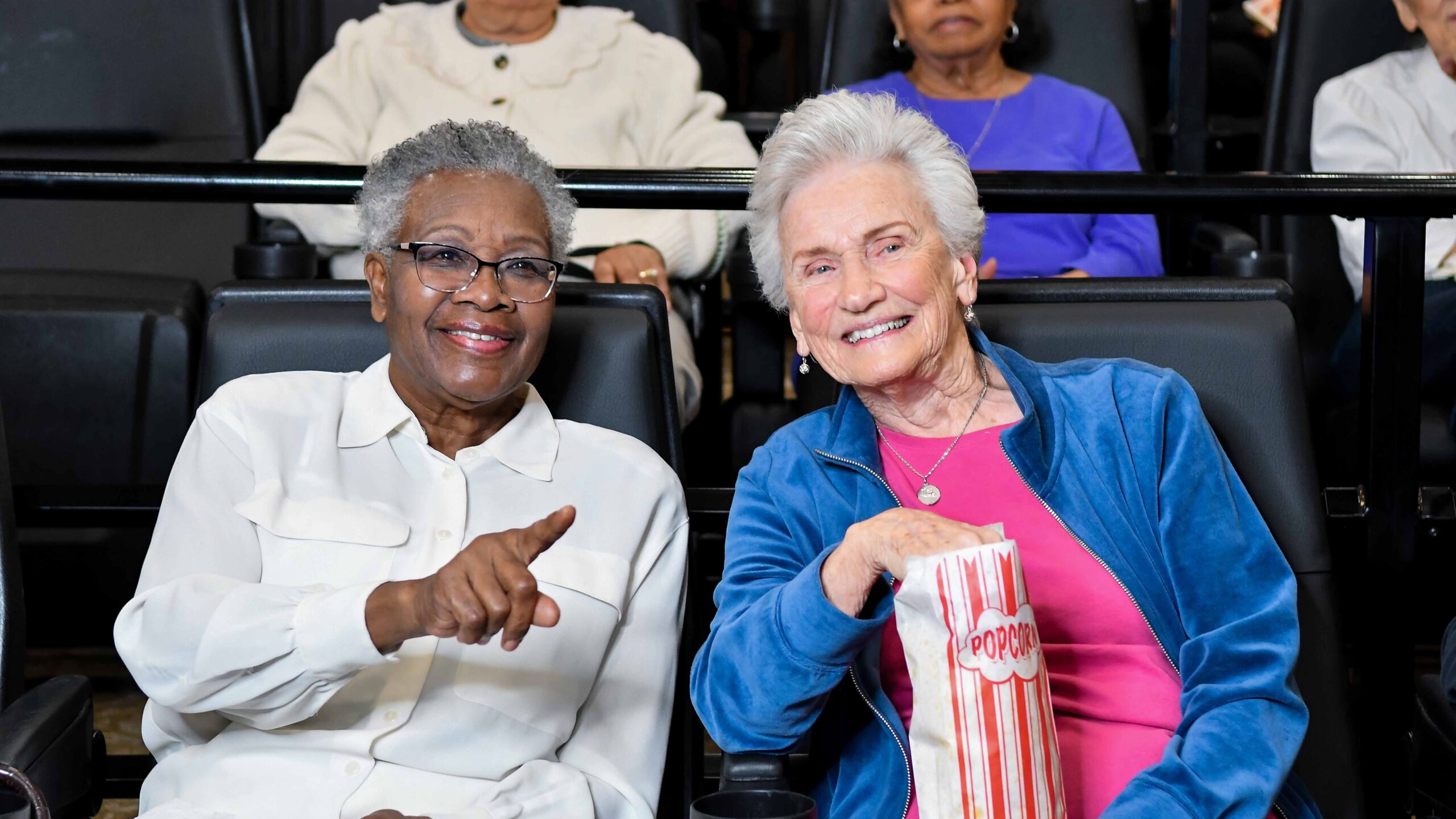 Two older women sitting in a movie theater, smiling. One is pointing and the other is holding a striped popcorn bag. Several other people are seated in the background.