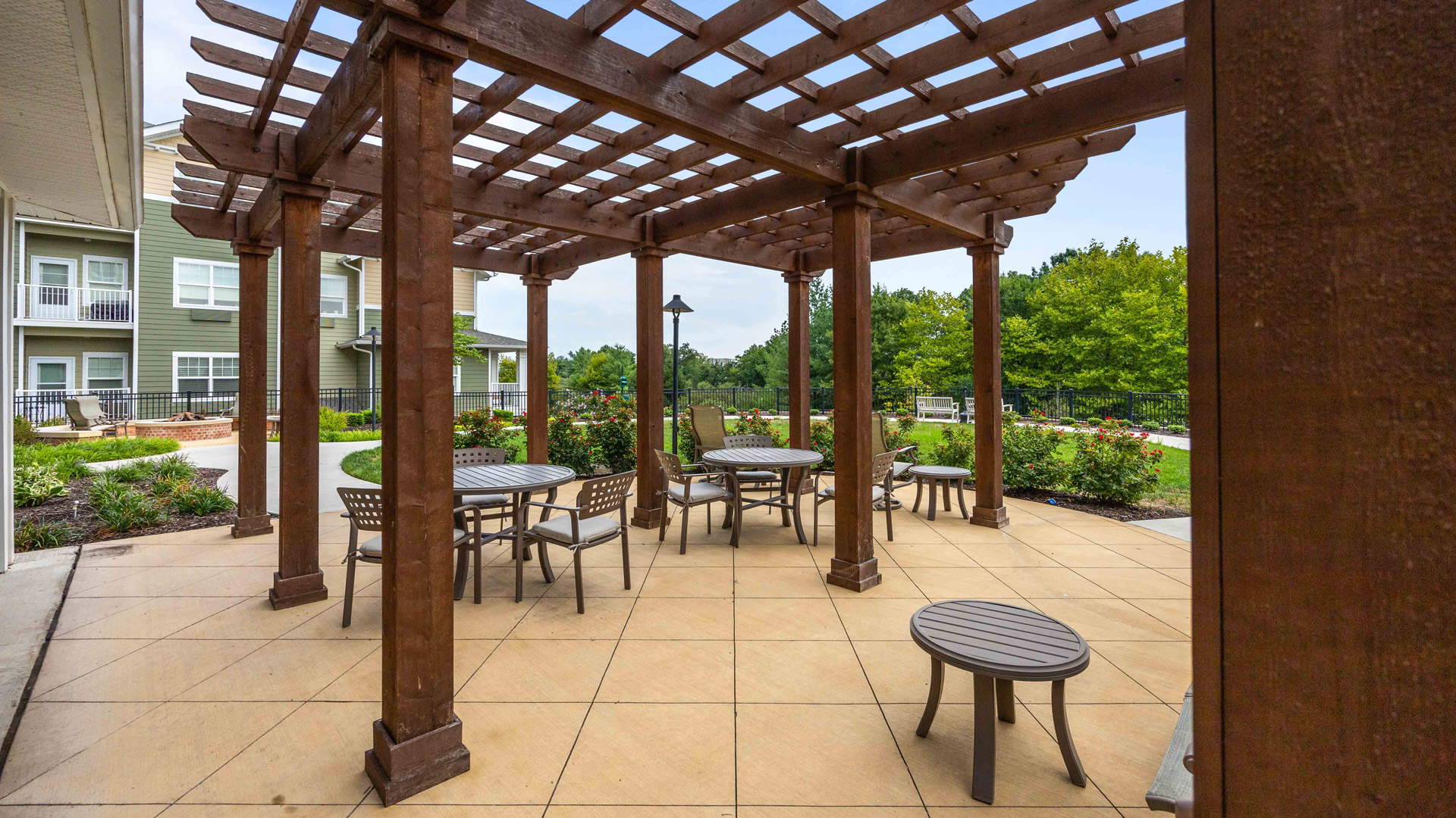 Outdoor patio area with a wooden pergola, tables, chairs, and surrounding greenery. The space is paved and situated next to a residential building with windows and balconies.
