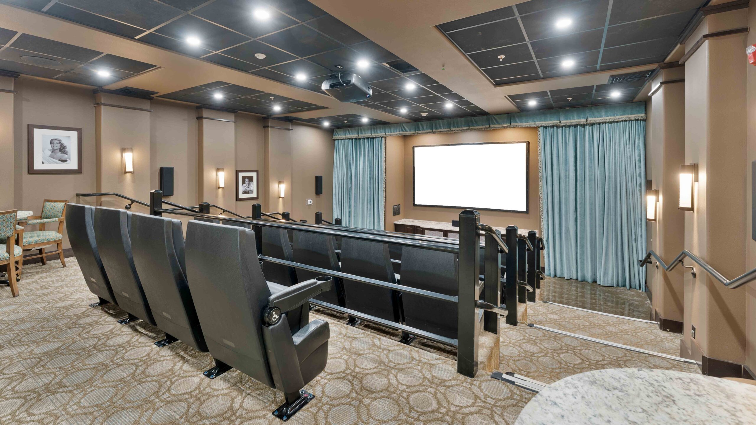 A modern home theater room with several rows of black recliner seats facing a large blank projector screen, soft lighting, framed wall art, blue curtains, and carpeted floor.