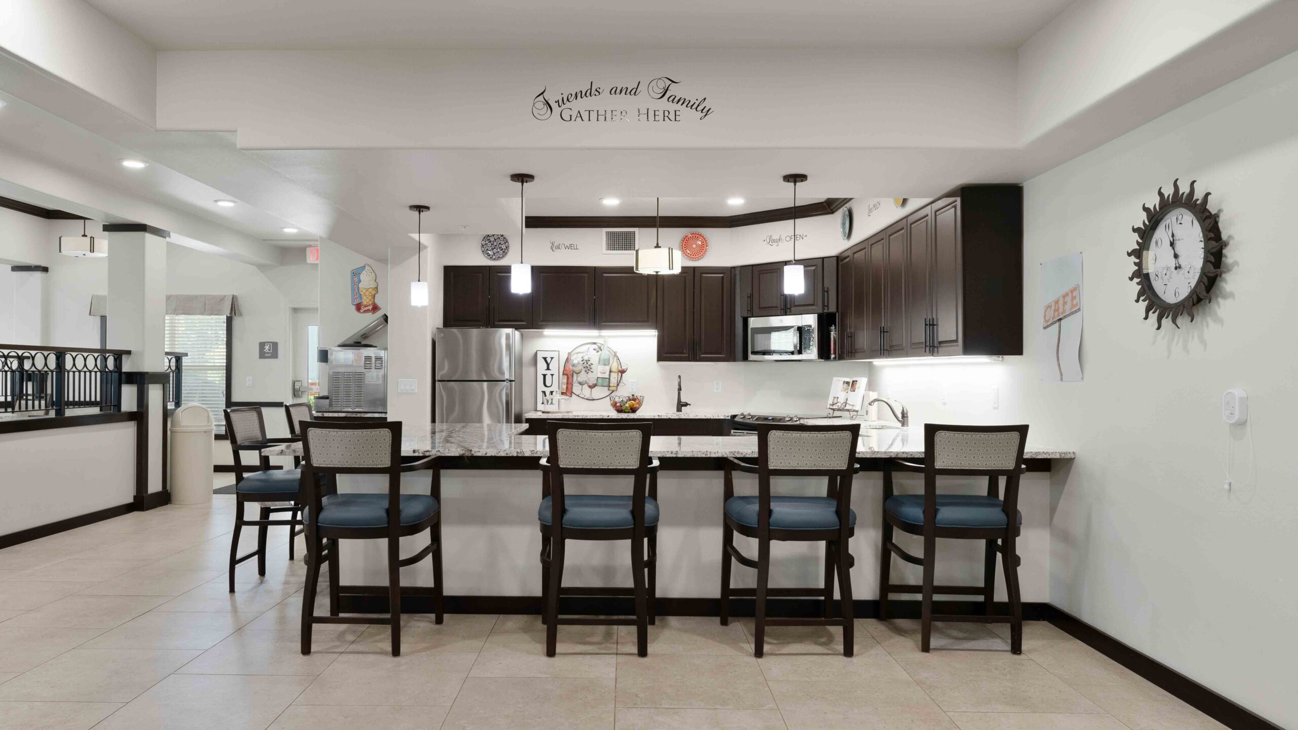 A modern kitchen with dark cabinets, stainless steel appliances, a marble island with five chairs, wall decor, and a large clock on the right wall. The phrase "Friends and Family gather here" is displayed above the kitchen.