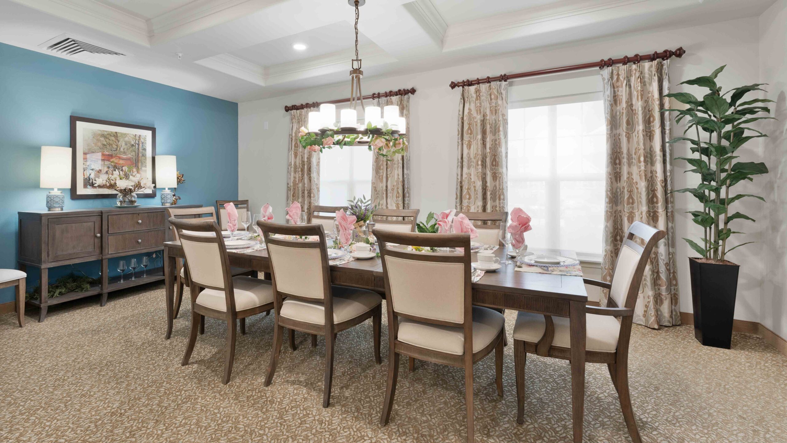 Elegant dining room with a long wooden table set for eight, beige cushioned chairs, a chandelier, large windows with patterned curtains, blue accent wall, sideboard, lamps, and a tall potted plant in the corner.