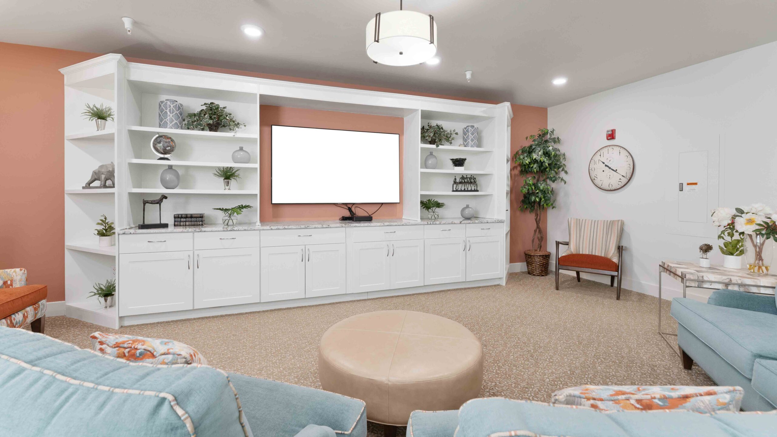 A bright living room with a large white entertainment center, a blank TV screen, shelves with decor and plants, pastel blue seating, a round beige ottoman, and a wall clock. The walls are light with one accent wall in peach.