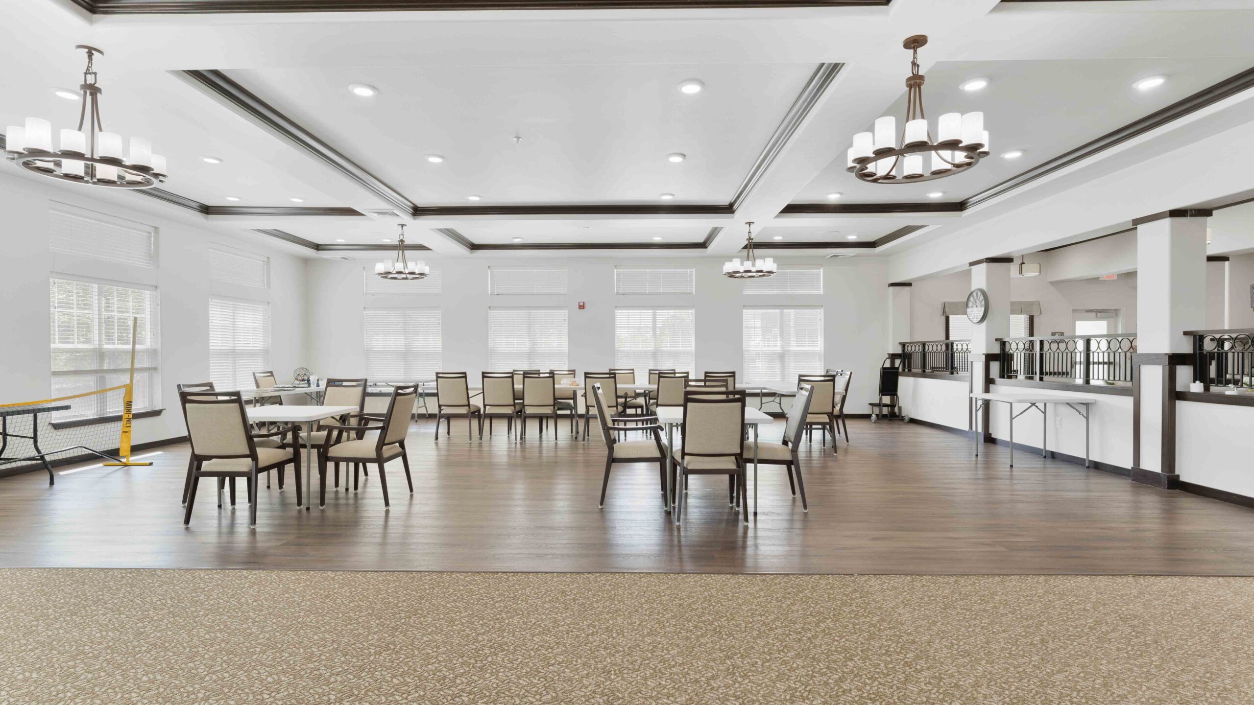 A spacious, well-lit dining area with several tables and chairs, elegant chandeliers, large windows, and wood flooring. The room appears clean and modern, with a buffet area in the background.