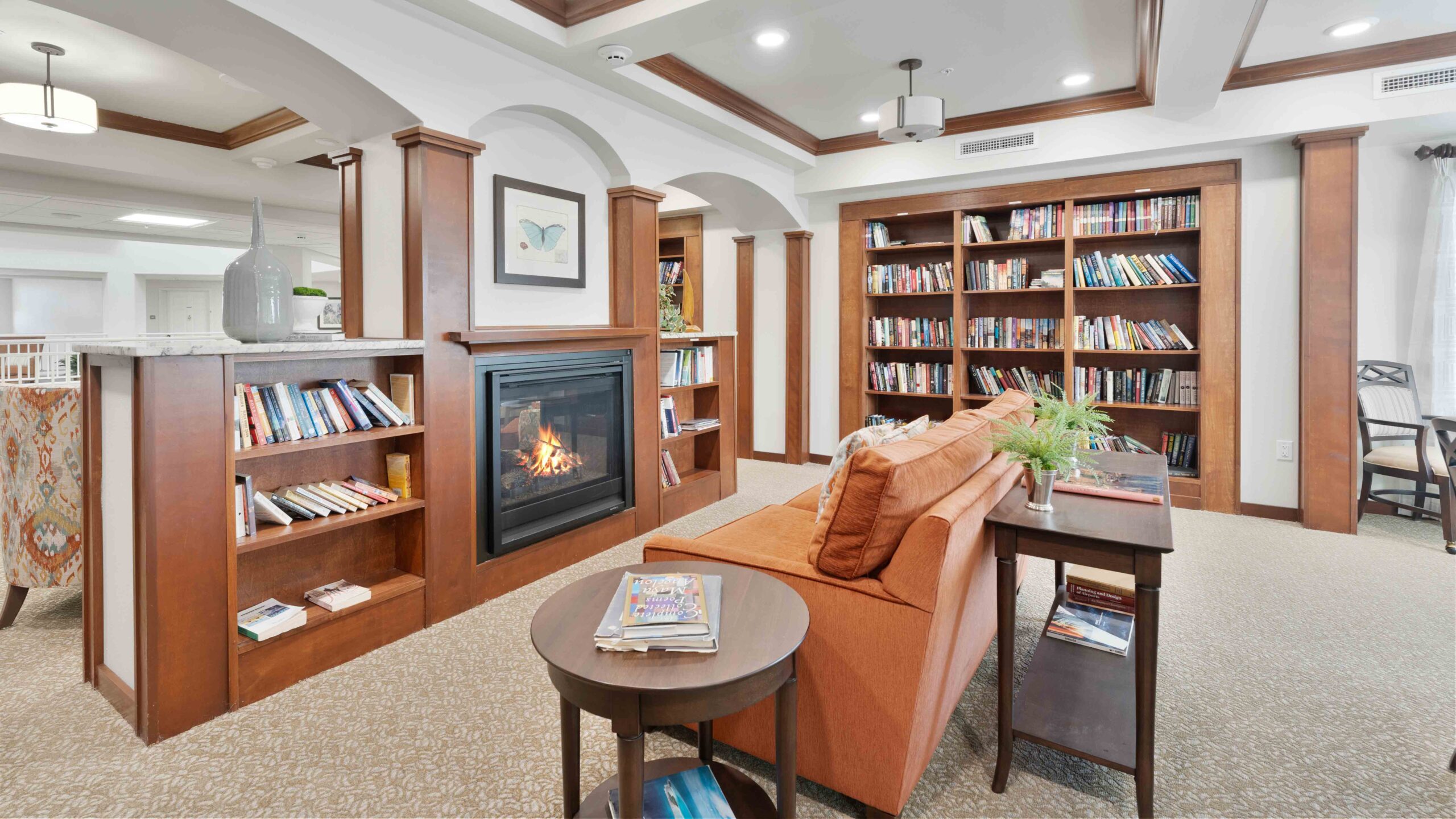 A cozy library room with wooden bookshelves, a lit fireplace, an orange sofa, and a small round table with magazines. Warm lighting and comfortable seating create an inviting reading space.
