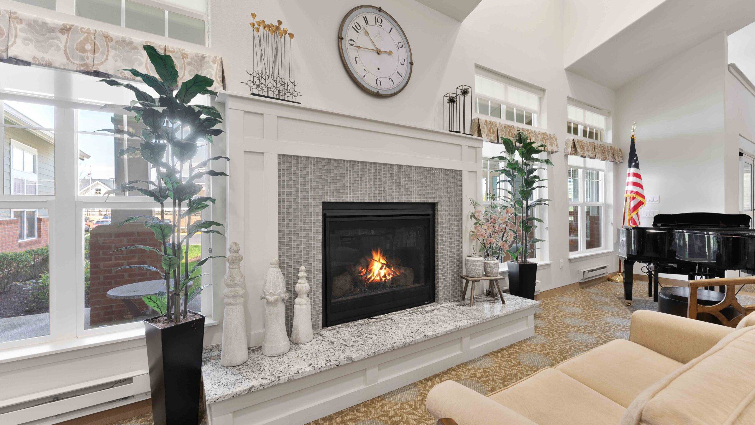 A cozy living room with a lit fireplace, decorative vases and sculptures on the mantle, large windows, an American flag, a black grand piano, and a beige sofa. A clock hangs above the fireplace.