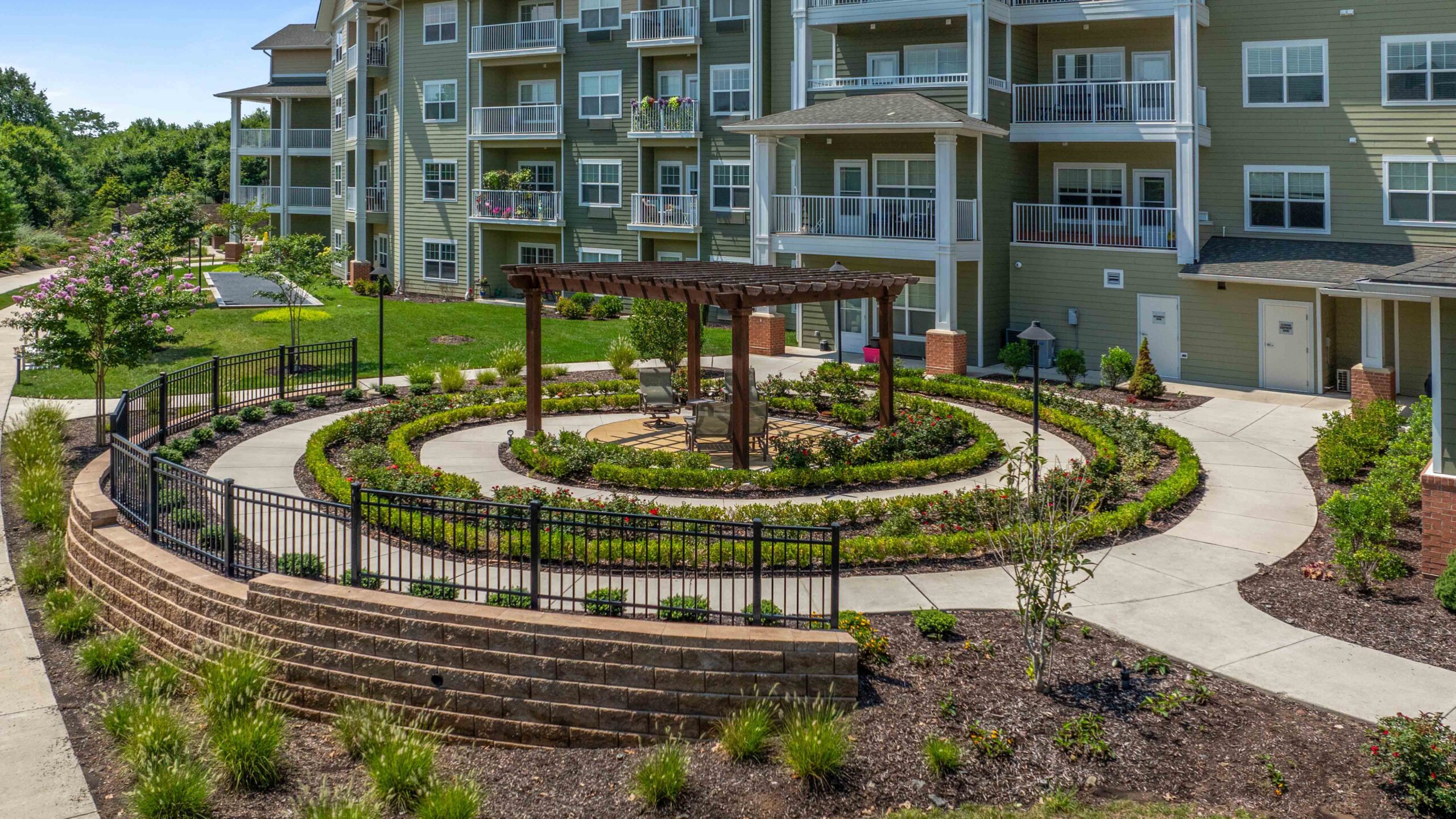 A landscaped courtyard with circular walking paths, a pergola with benches in the center, green plants, and a multi-story apartment building in the background.