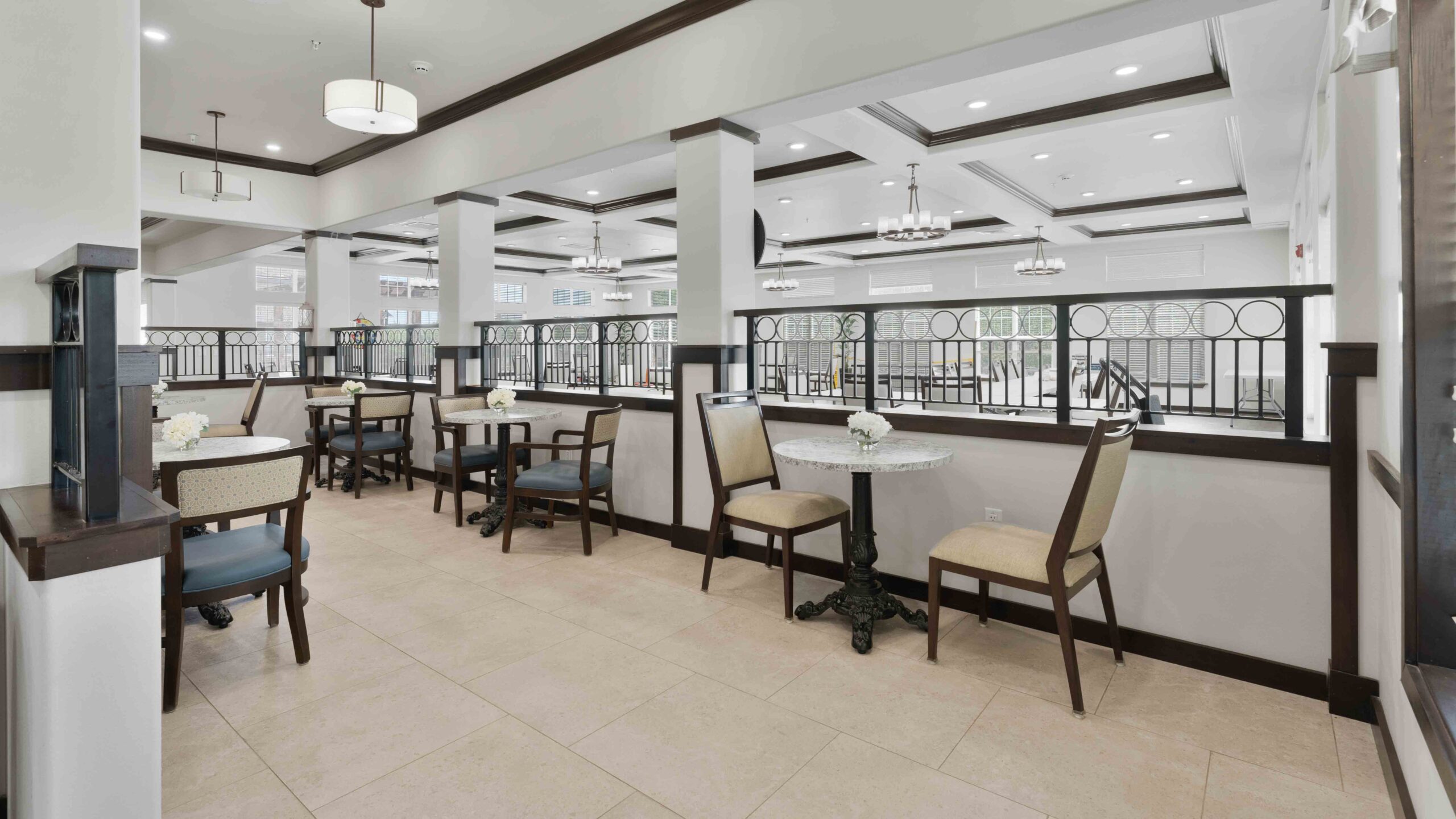 A bright, modern dining area with several small tables and chairs, neutral walls, large windows, and elegant light fixtures. The space has light tile flooring and decorative railing separating sections.