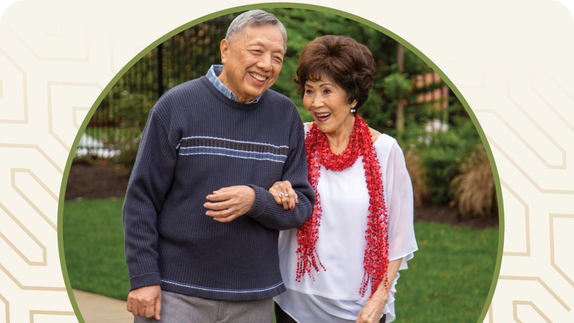 An elderly couple smiling and walking outdoors. The man, wearing a dark sweater, holds the arm of the woman in a white top with a red scarf. They are in a garden setting with greenery in the background.
