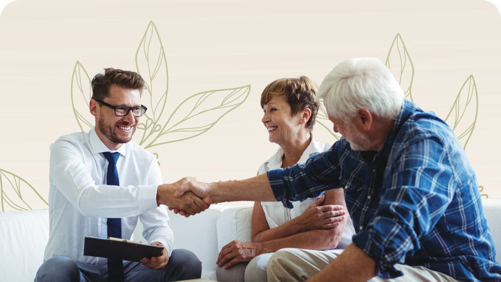 A man in a white shirt and tie shakes hands with an older man in a plaid shirt. An older woman, sitting next to the older man, smiles. They are seated in front of a light background with leaf patterns.