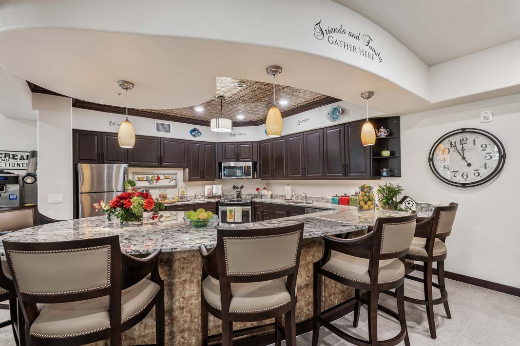 A spacious, modern kitchen featuring a large marble island with six cushioned bar chairs. Dark wood cabinetry lines the walls, and pendant lights hang from the ceiling. The decor includes flowers, a large clock, and a sign that reads 