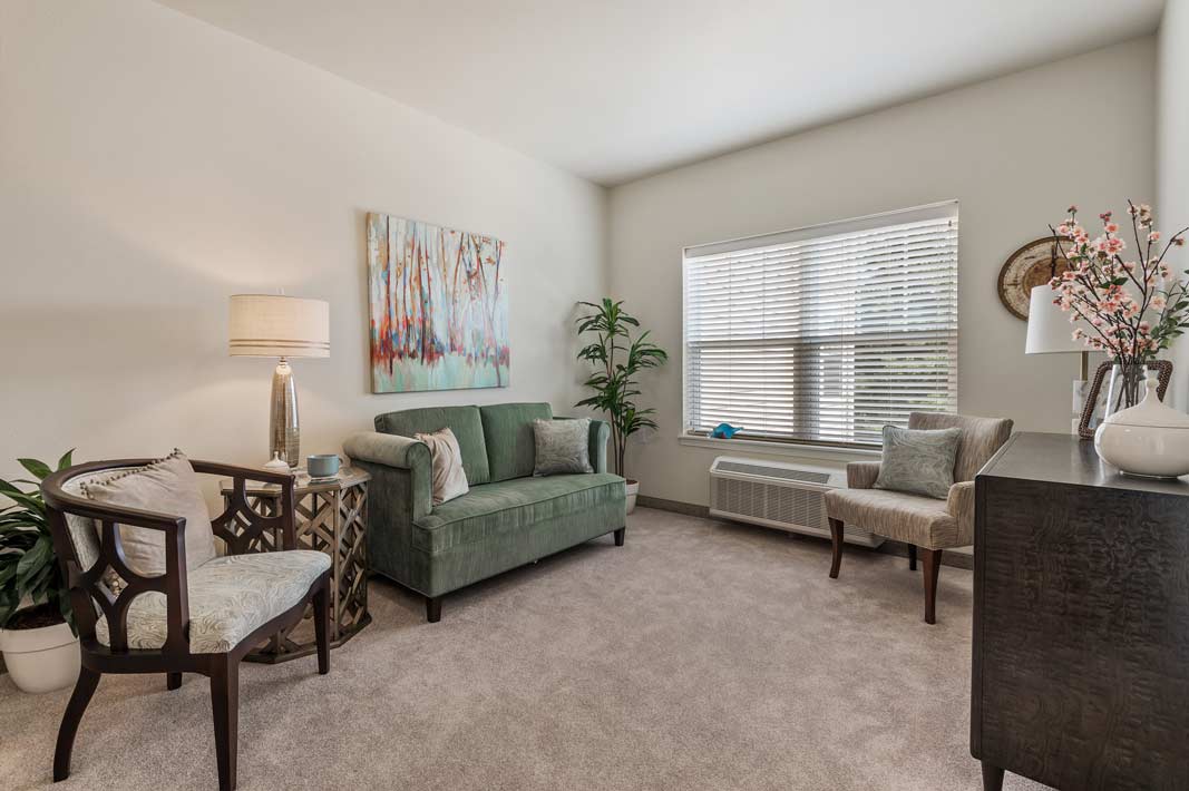 A cozy living room with light beige carpet and off-white walls, featuring a green sofa, two stylish chairs, a wooden coffee table, and a framed abstract painting. The room is adorned with indoor plants, a table lamp, and a window with blinds letting in natural light.